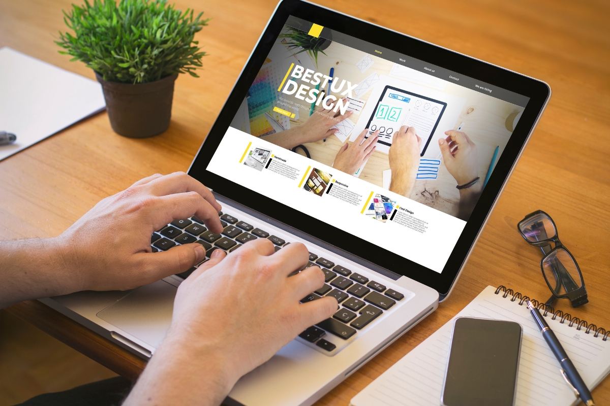 Person using a laptop displaying a website about ux design created with Elementor in WordPress on a wooden desk with a notepad, glasses, and a small potted plant.