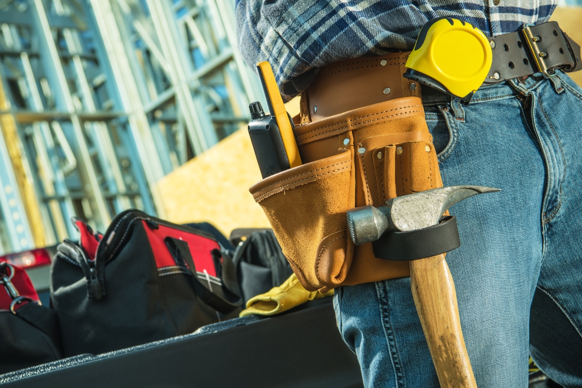 Construction worker with a Houzz Pro tool belt carrying a hammer, standing near a tool bag in a construction site.