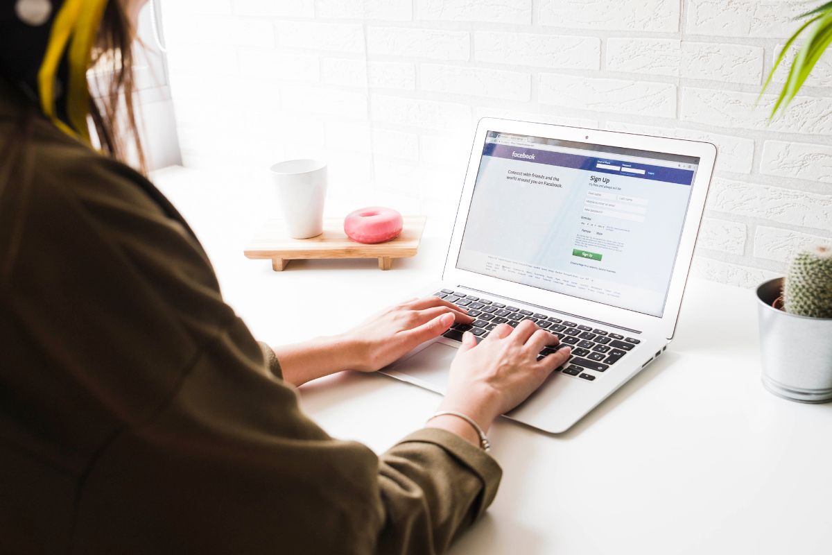 A woman wearing headphones using a laptop with a "how to create a Facebook business account" guide, sitting at a white desk with a coffee cup, apple, and potted plant.