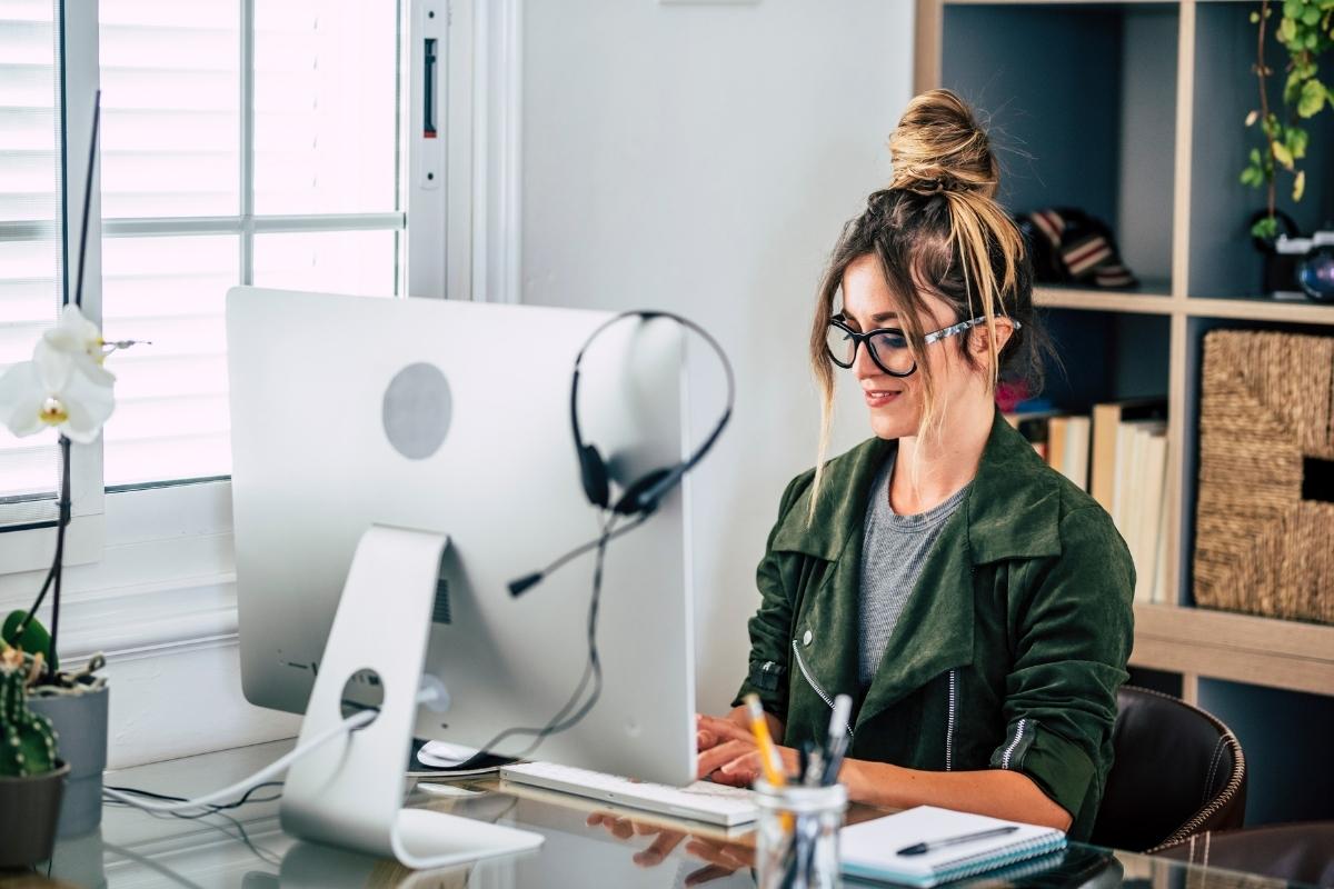 A woman wearing glasses is working at a desk with an iMac and a podcasting microphone, focused on web design and SEO.