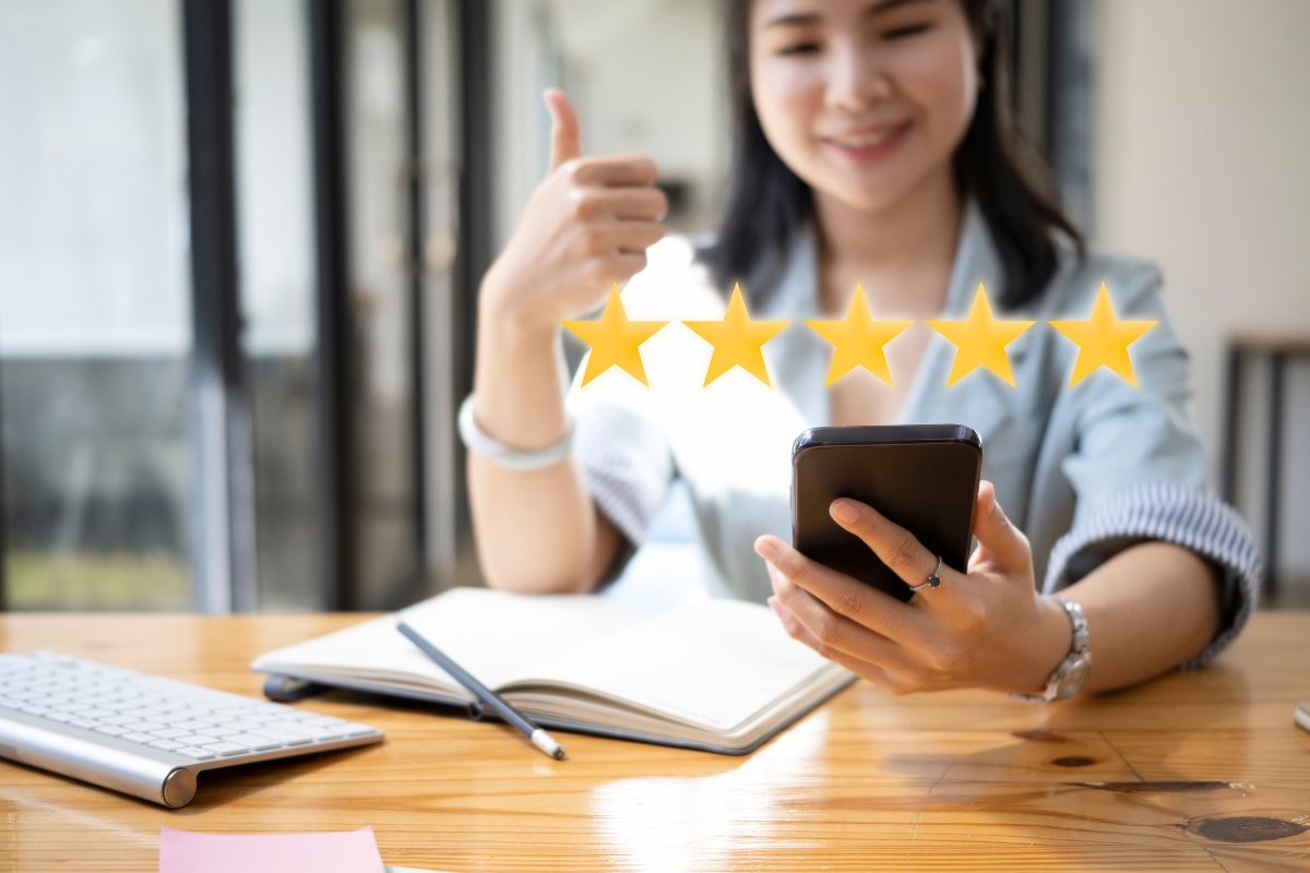 A woman sitting at a desk, giving a thumbs up with one hand and holding a smartphone in the other, with five stars displayed above the phone, indicating a positive online reputation.