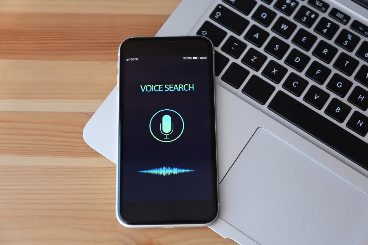 A smartphone with voice search capability next to a laptop on a wooden table.