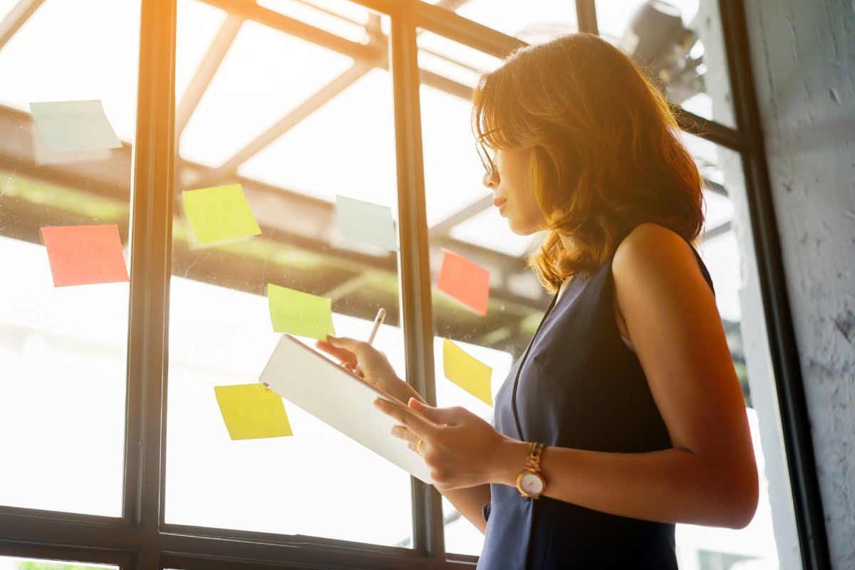 Businesswoman fixing suspended business profiles while writing on sticky notes in front of a window.