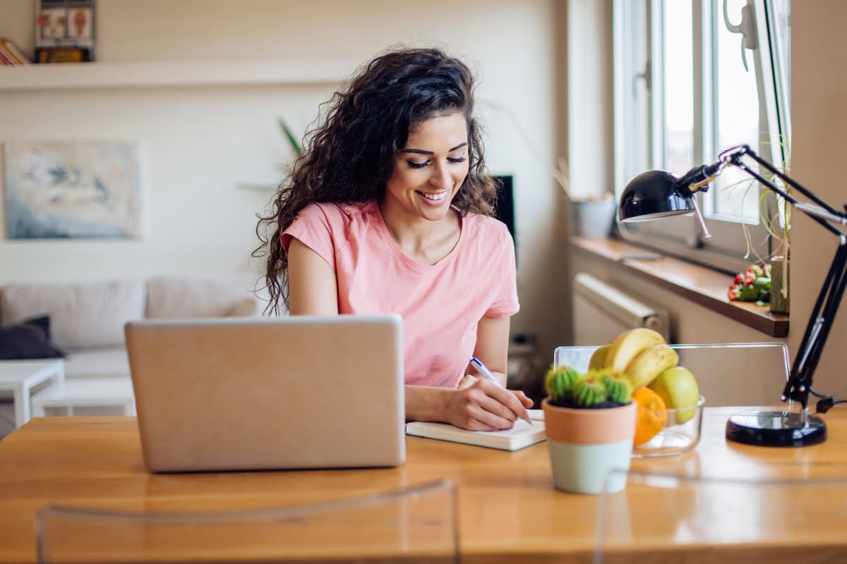 A woman sitting at a table with a laptop, managing her home improvement business while enjoying some fruit.