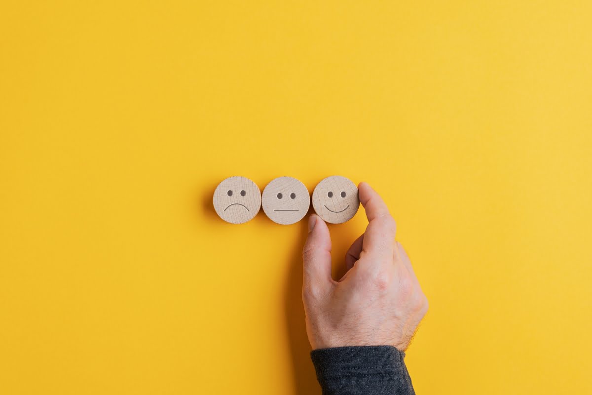 A man's hand is holding three wooden smiley faces on a yellow background.