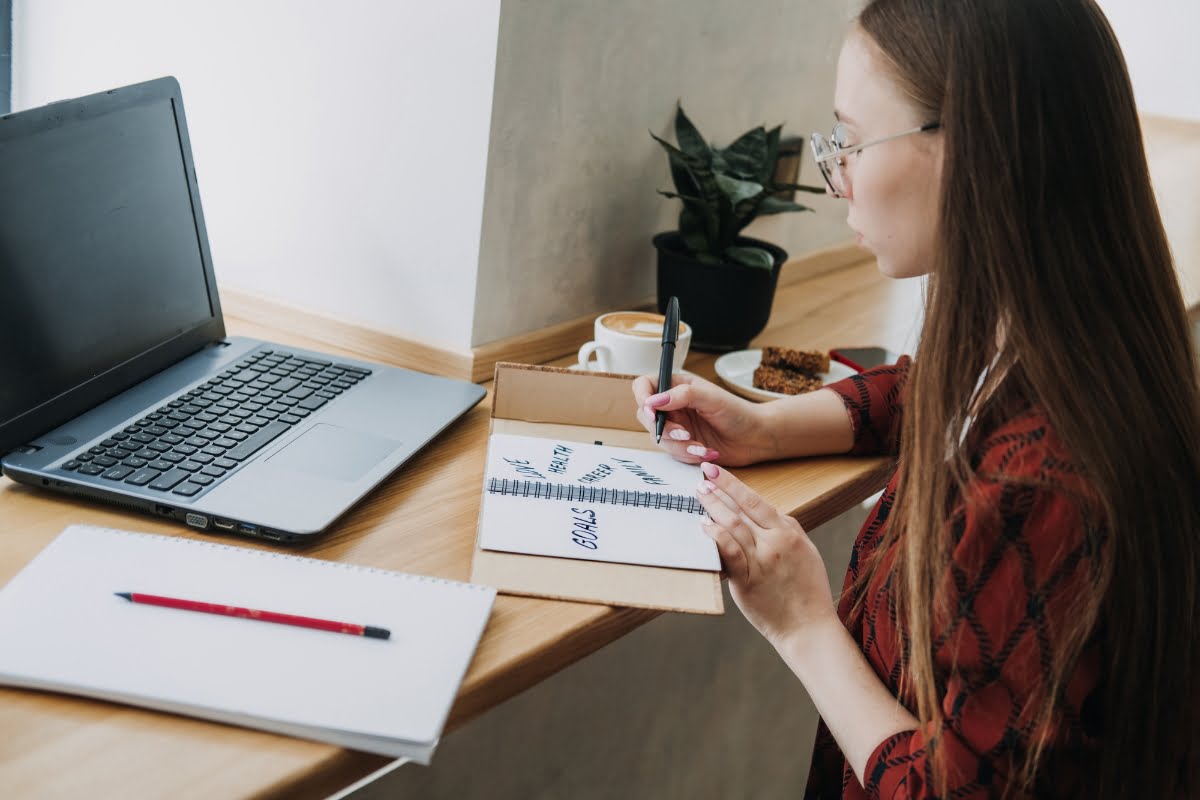 A woman, working from home, diligently writes on a notebook while sitting in front of her laptop.