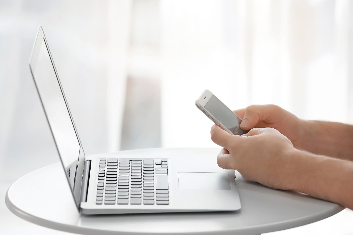 A man is using a cell phone while sitting at a table with a laptop, possibly researching how to update WordPress.