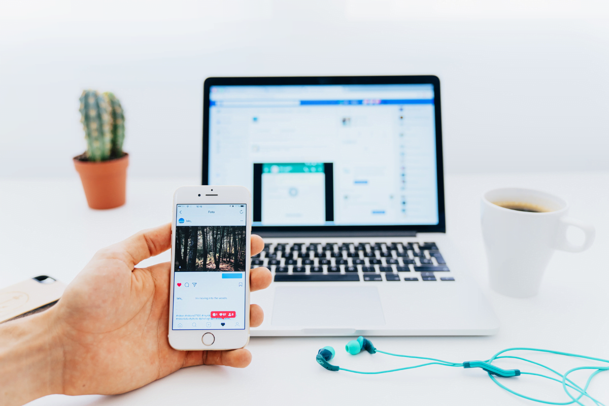 A hand holding a cell phone next to a laptop showing Instagram on the screen.