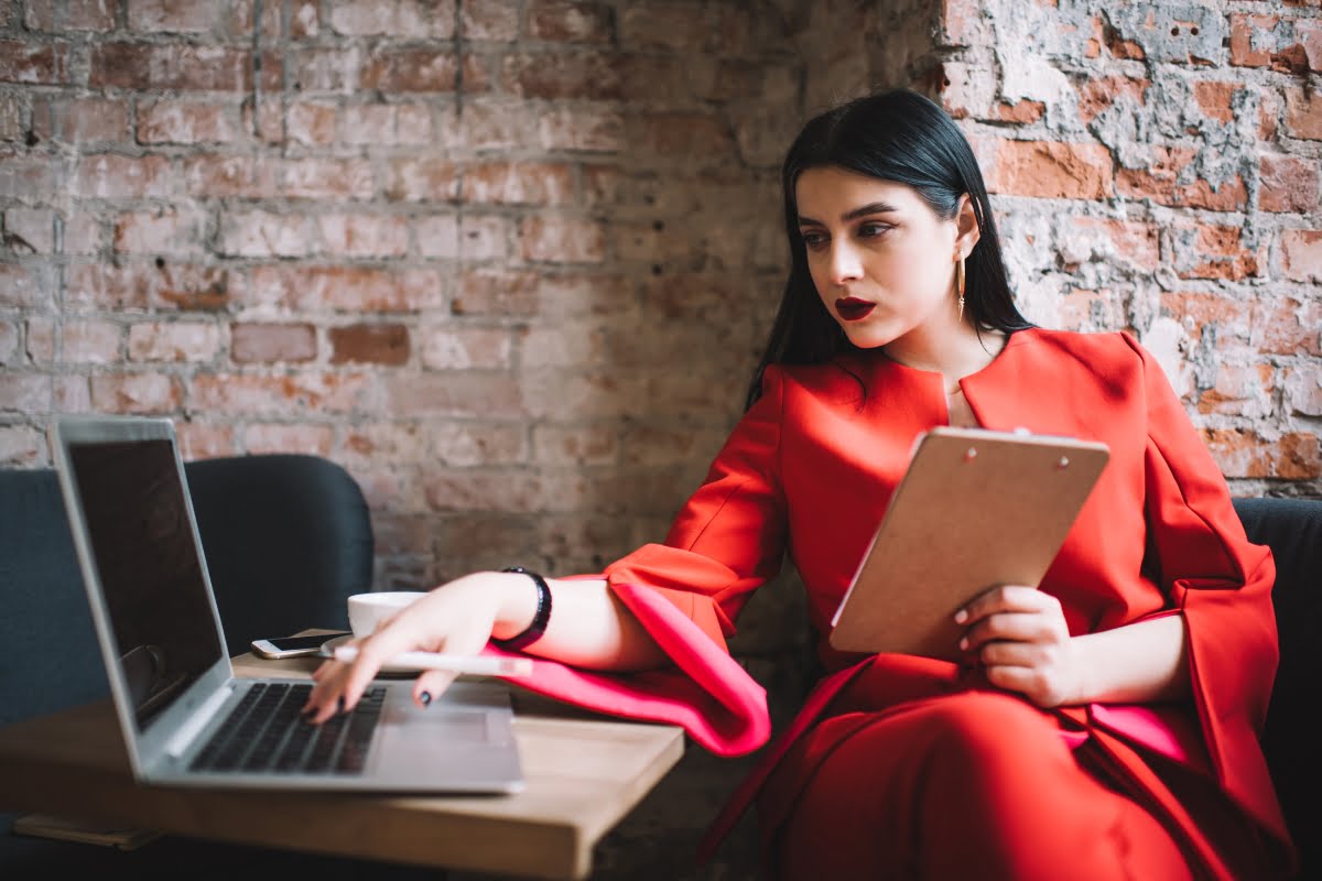 A woman in a red dress is writing copy on a laptop in a coffee shop.