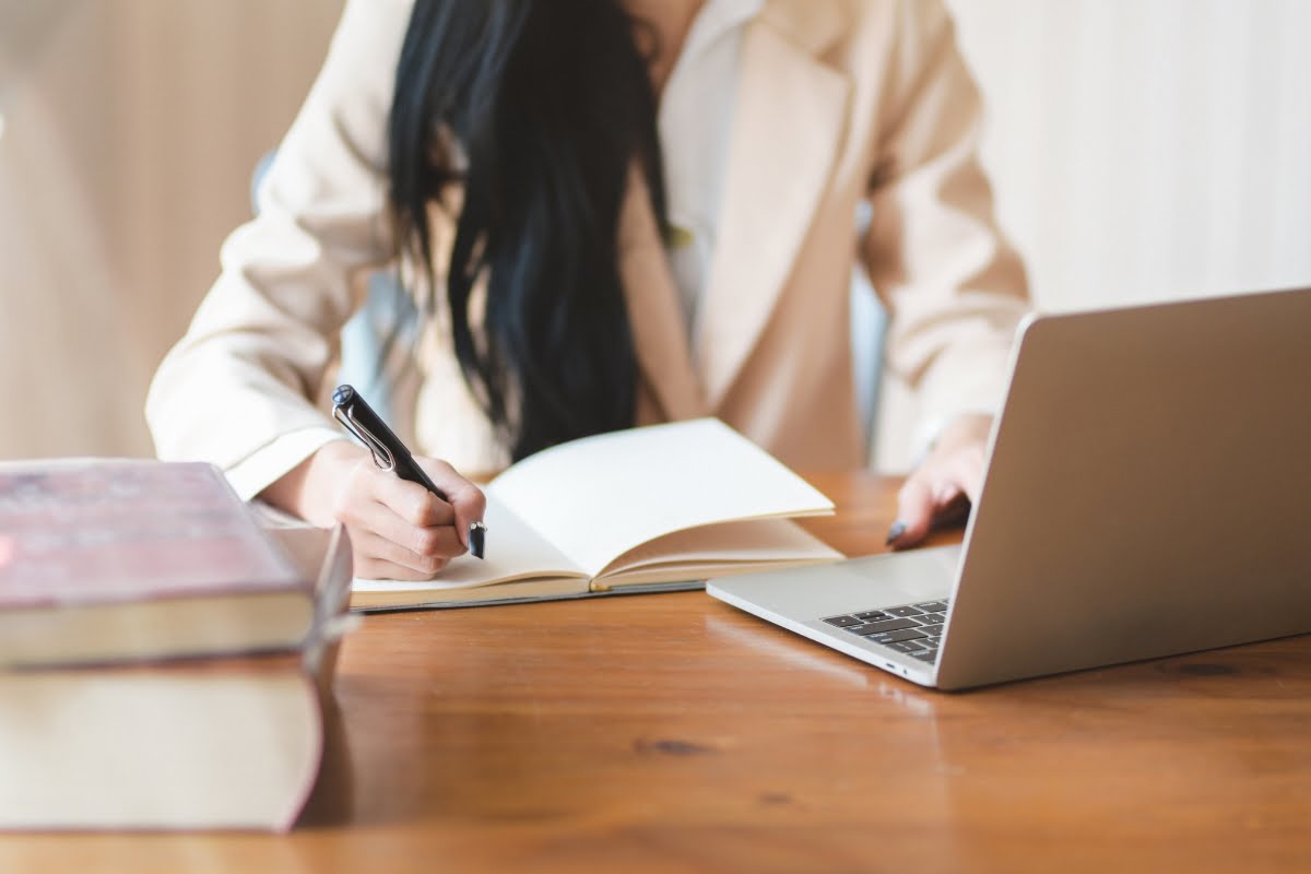 A woman writing copy at a desk with a laptop and a notebook.