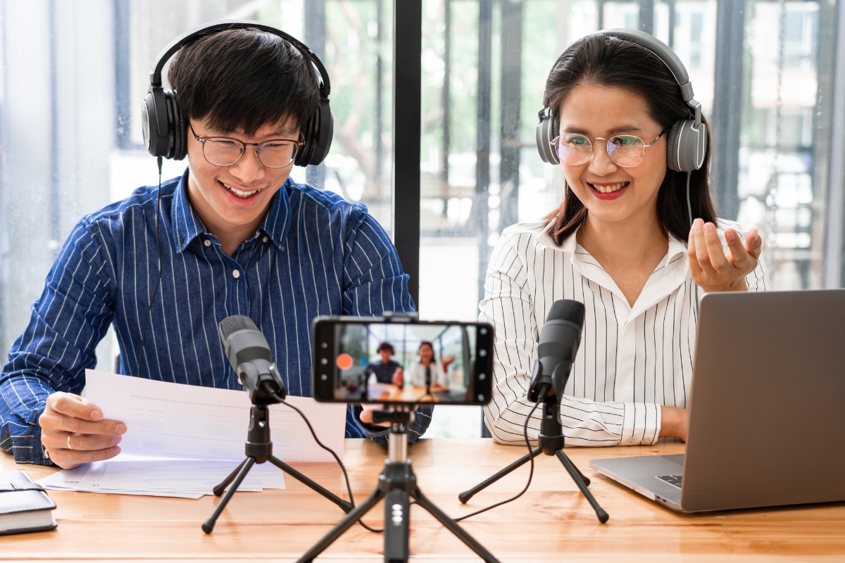 Two asian people sitting at a desk, engaged in creative content ideas with headphones and a laptop.