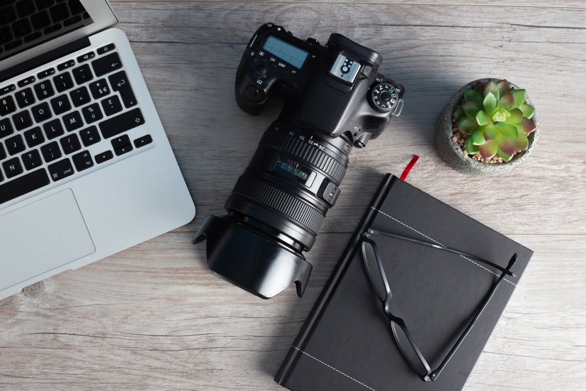 An evergreen laptop, camera, notebook and cactus on a wooden table.