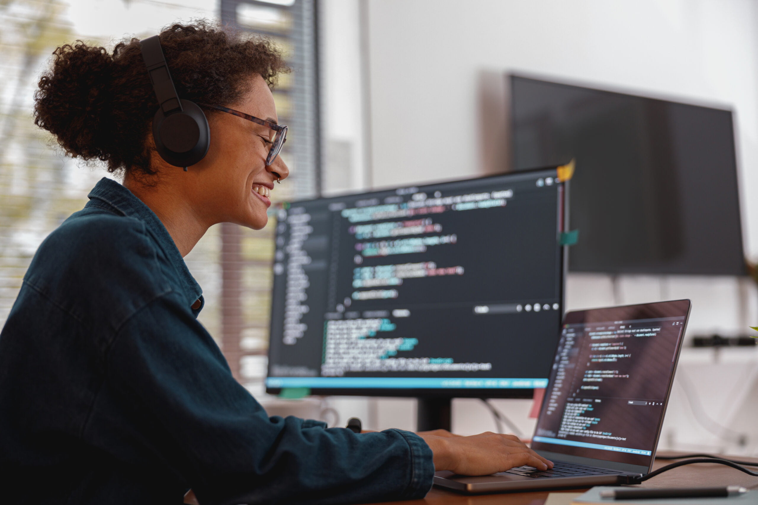 A woman wearing headphones is working on a laptop in front of a computer screen.