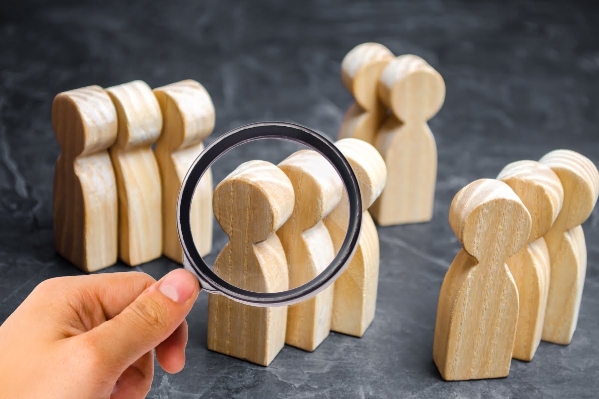 A hand holding a magnifying glass over a group of wooden figures, showcasing the potential for targeted spa email marketing.