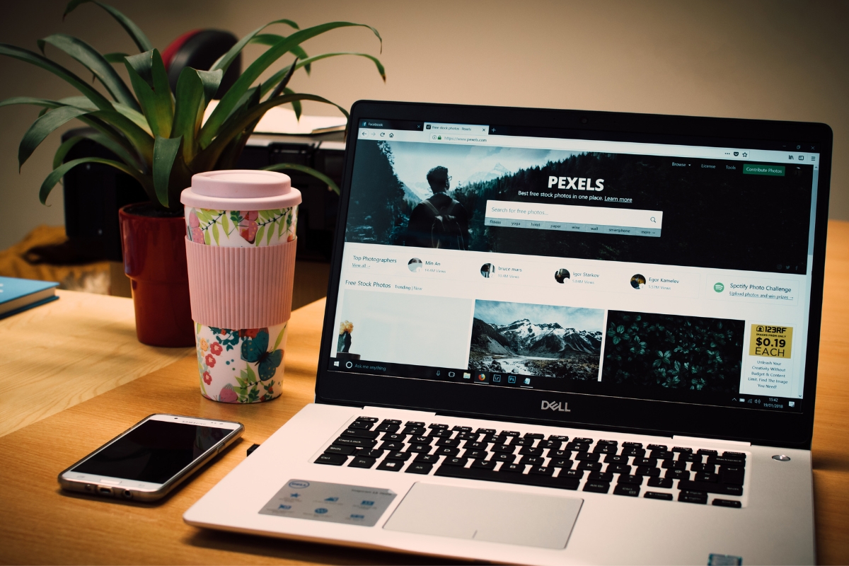 A laptop is sitting on a desk next to a cup of coffee and a plant in website photos.