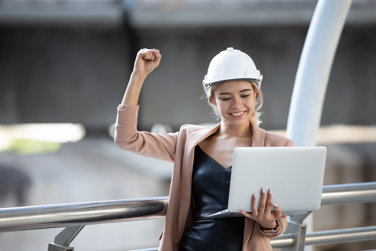 A woman wearing a hard hat and holding a laptop with her hand raised as a sign of success.