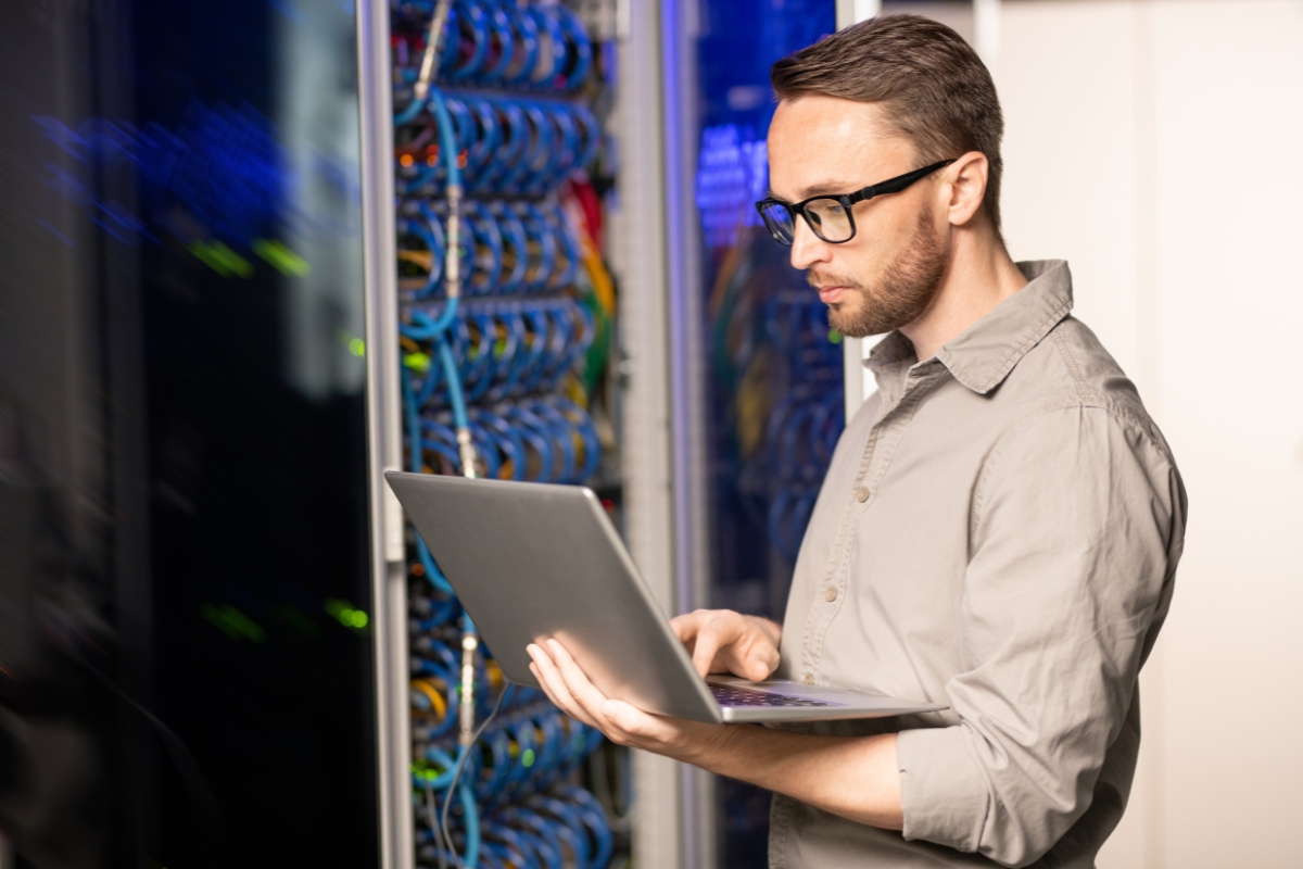 A man in glasses is utilizing a laptop in a server room to assist with WordPress.