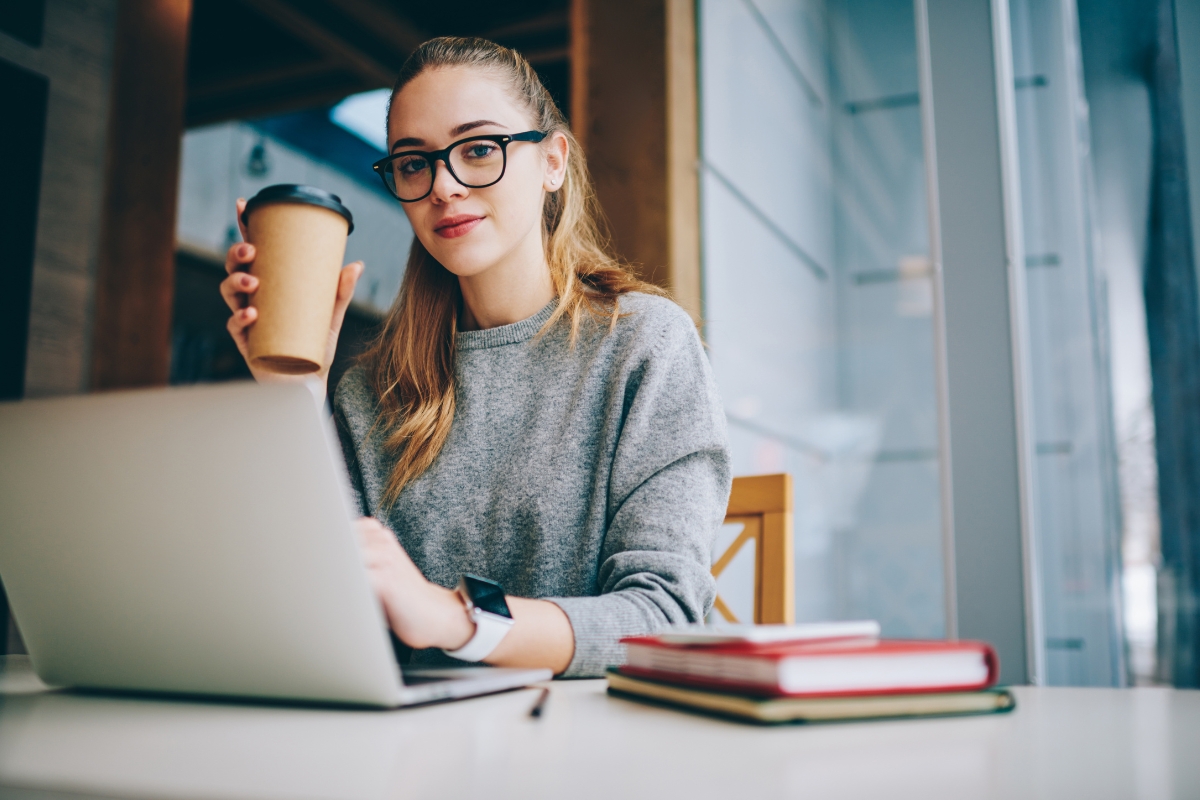 A woman in glasses is sitting at a table with a laptop and a cup of coffee, working on her seo copywriting.