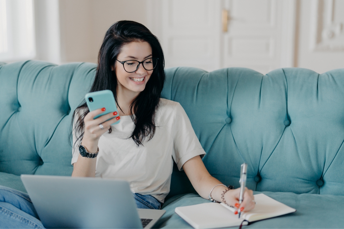 A woman sitting on a couch with a laptop and cell phone, multitasking for work.