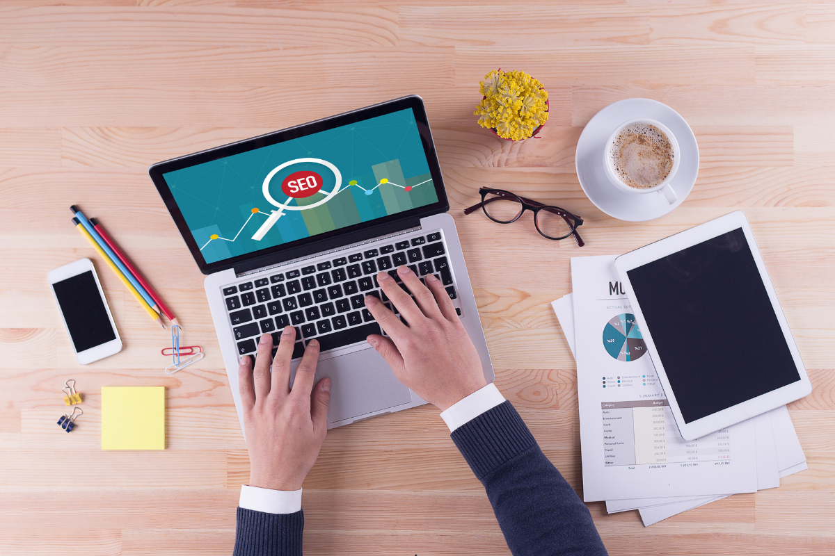 A man's hands typing on a laptop with a magnifying glass and the word SEO on a wooden office desk.