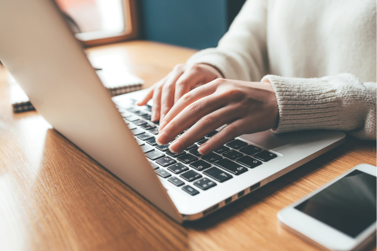 A woman typing a website copy on a laptop with a cellphone nearby.