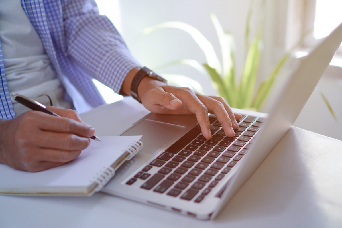 A man typing on a laptop while holding a pen, researching the purpose of a website.