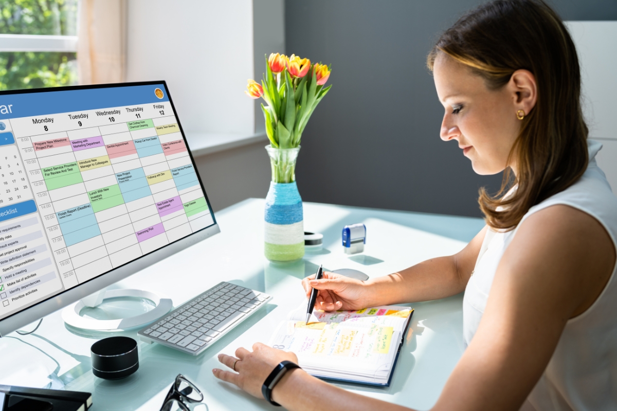 A woman taking notes from a content calendar on her computer screen in a bright office setting.