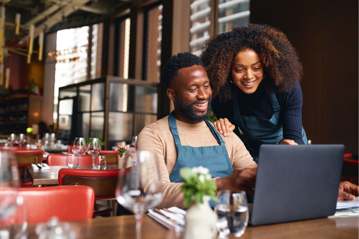 Restaurant owners looking at a laptop together with smiles in an empty dining area, reviewing a Google business ownership request.