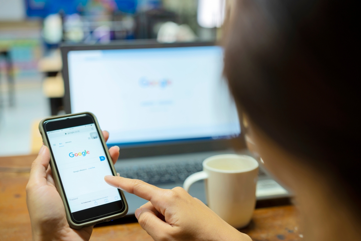 Person using Google Guaranteed local services search on a smartphone with a laptop displaying Google's homepage in the background.