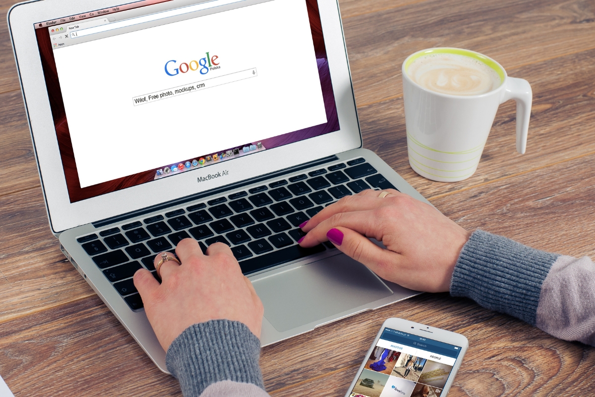 Person using Google Guaranteed local services search on a MacBook with a smartphone and a cup of coffee on the desk.