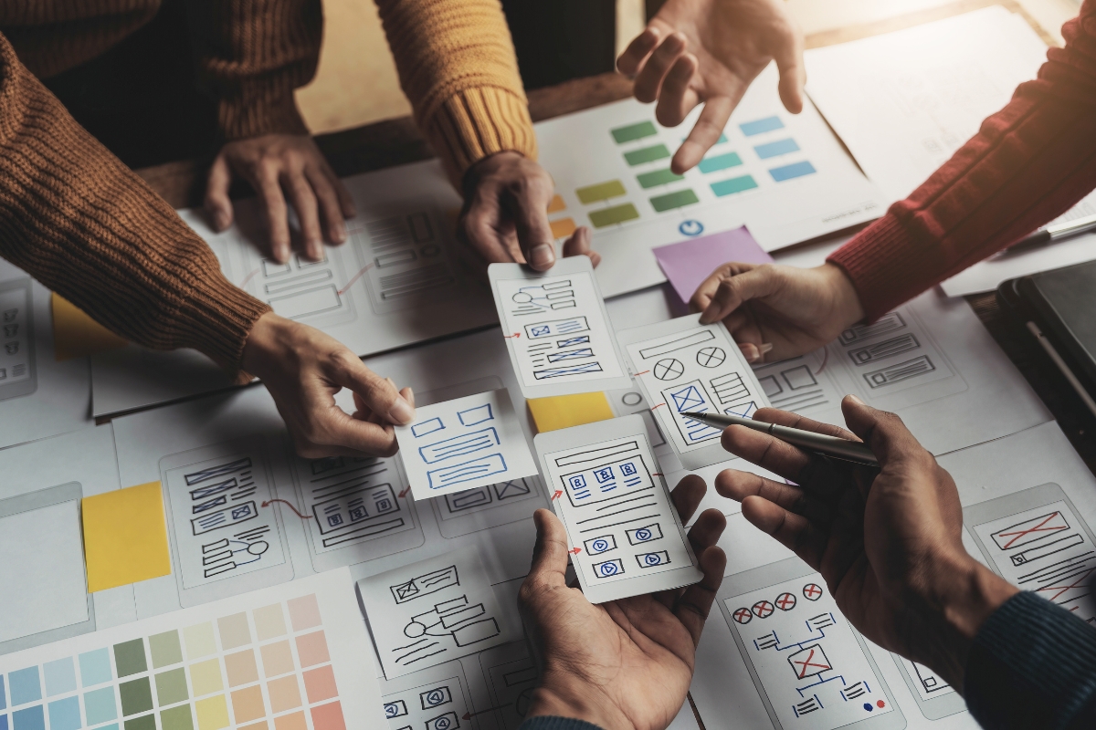 Team members collaborating on how to create wireframes for a user interface design with paper sketches and color palettes on a table.