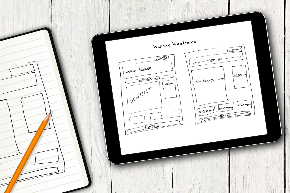 A tablet displaying how to create wireframes sketches next to a paper notepad and pencil on a wooden surface.