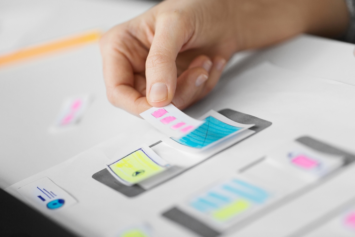 A person's hand placing a pink sticky note on printouts of mobile interface designs to explain how to create wireframes.