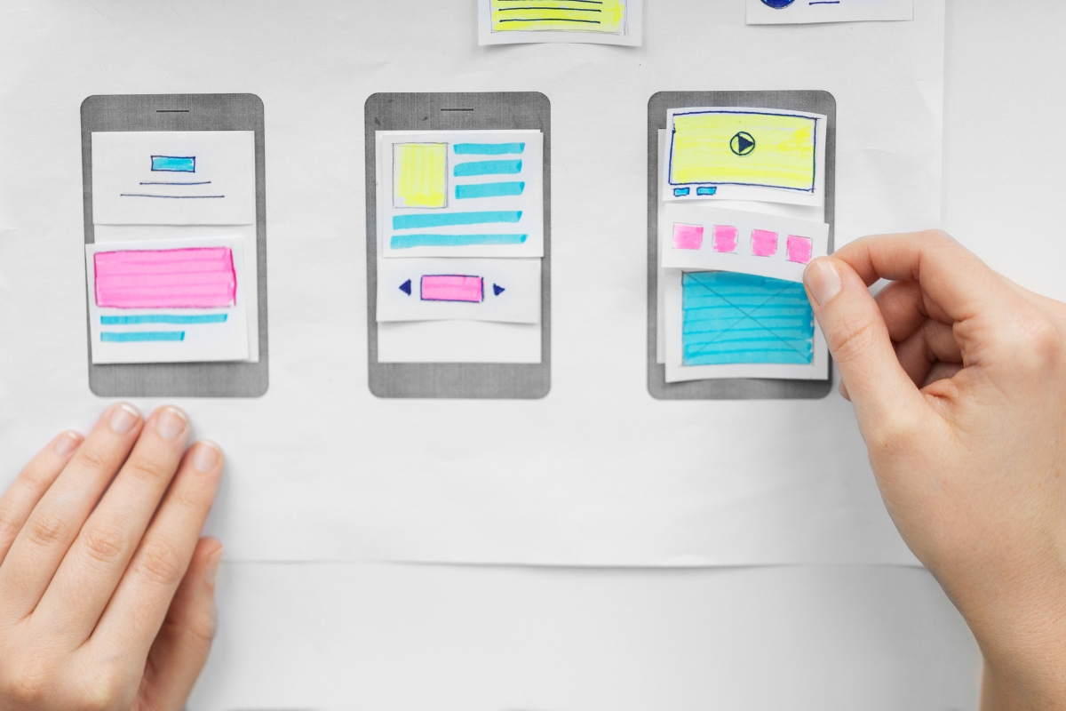 A person's hands arranging paper sketches of how to create wireframes for mobile app interfaces on a whiteboard.