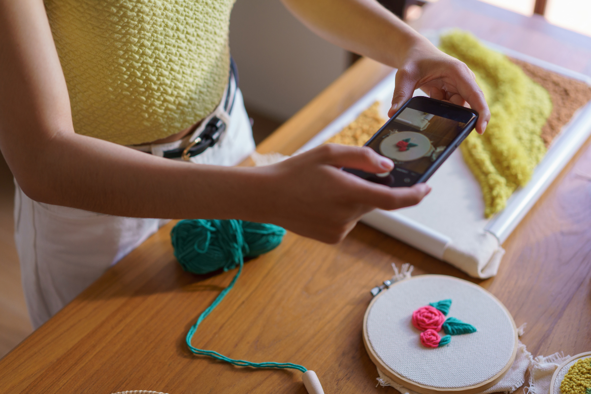 A woman is using a cell phone to take a picture of a crocheted hoop for her small business.