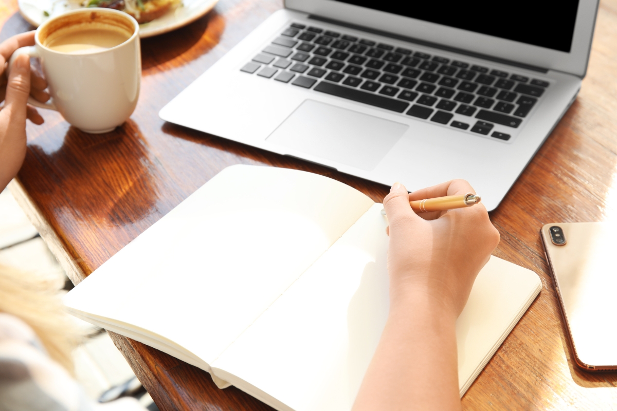 A woman crafting her medium SEO strategy in a notebook with a cup of coffee and a laptop.