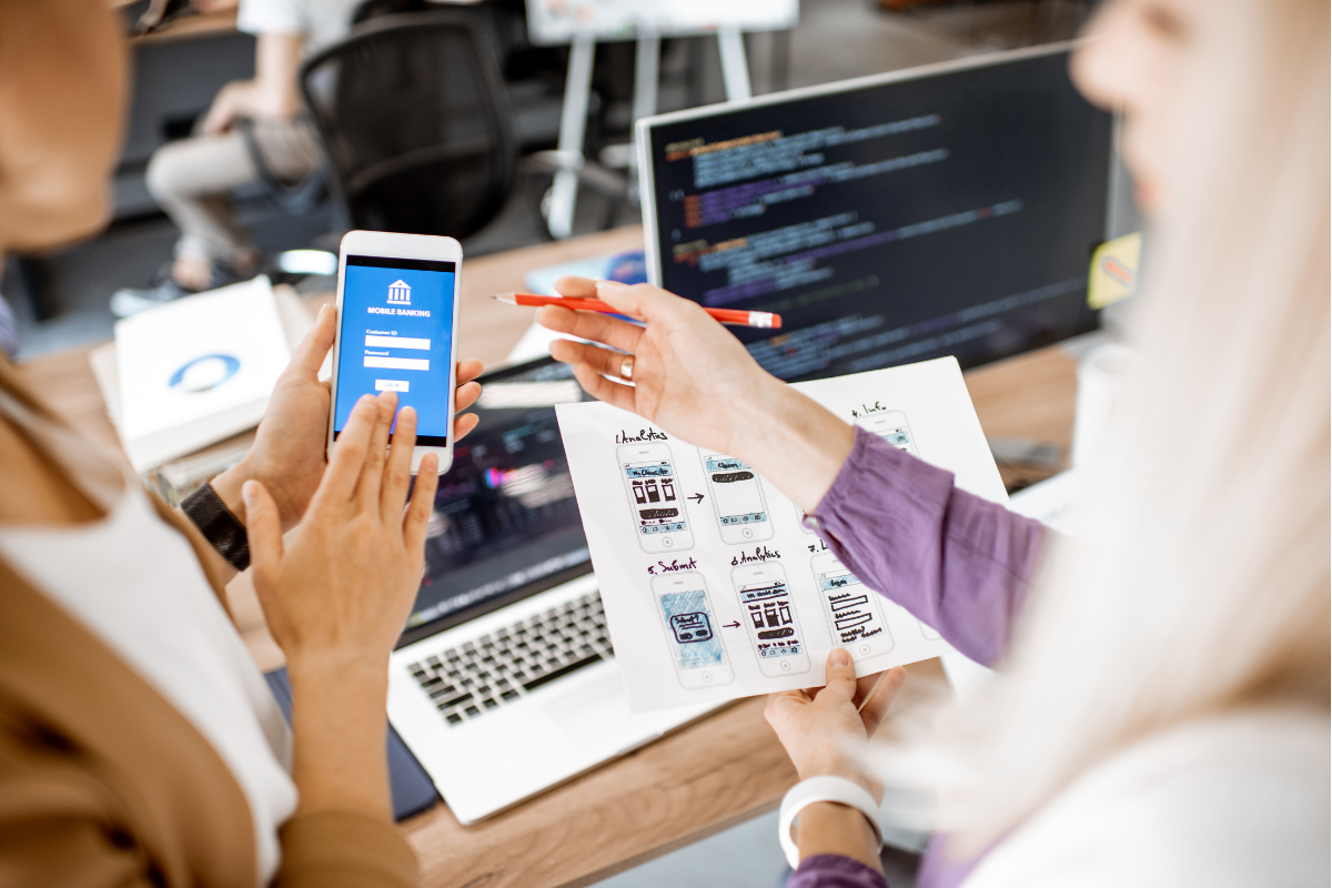 Two women looking at a computer screen and a smartphone to check on mobile responsiveness.