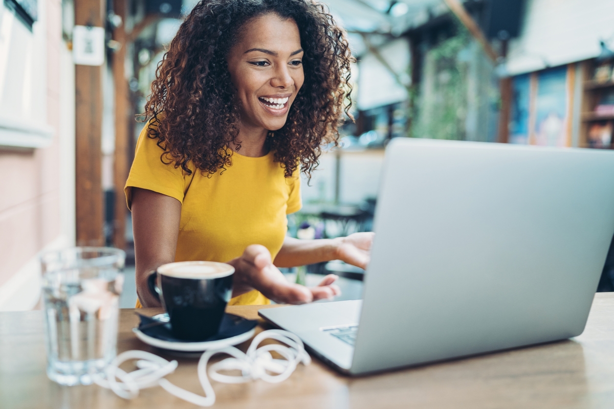 A woman in a yellow top smiling and gesturing during a video call on her laptop at a cafe table with a coffee cup and glass of water, discussing Reddit marketing strategies.