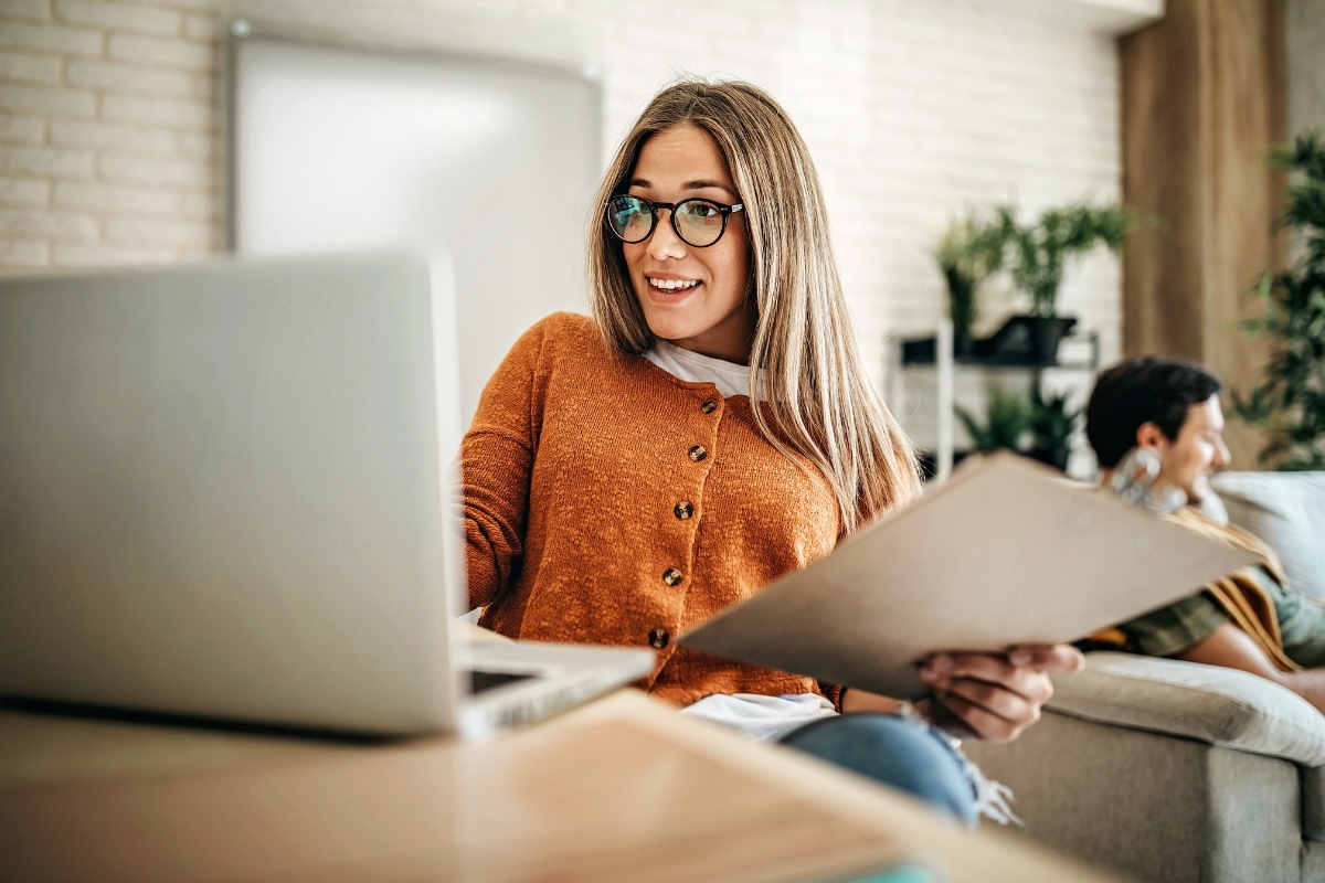 Woman in glasses working on laptop and holding documents related to Reddit marketing, with another person in the background.