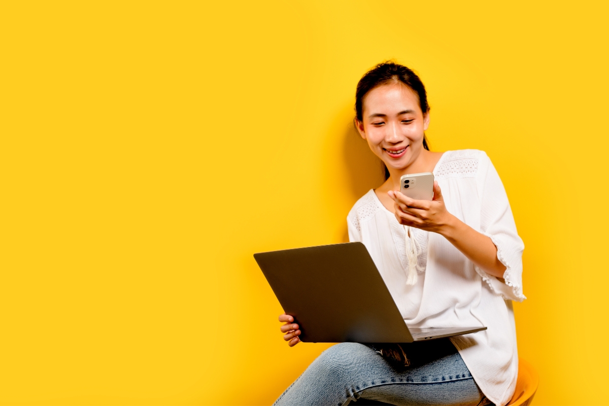 Woman smiling while using a smartphone and laptop for Reddit marketing against a yellow background.