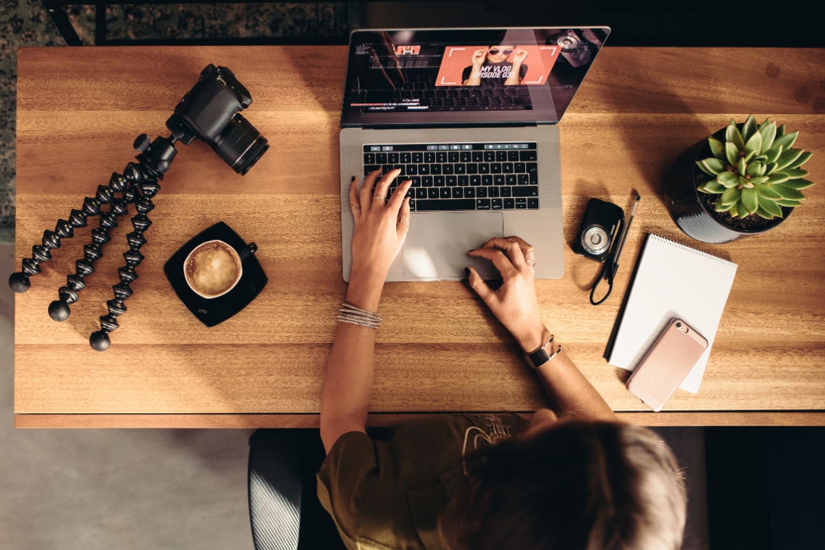 Person working on a laptop at a wooden desk with photography equipment, a cup of coffee, and reddit marketing materials.