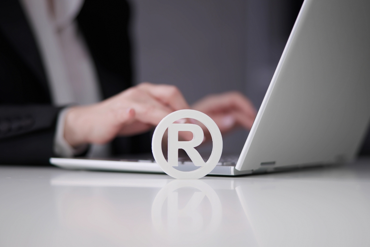 A registered trademark symbols standing on a table with a person in business attire working on a laptop in the background.