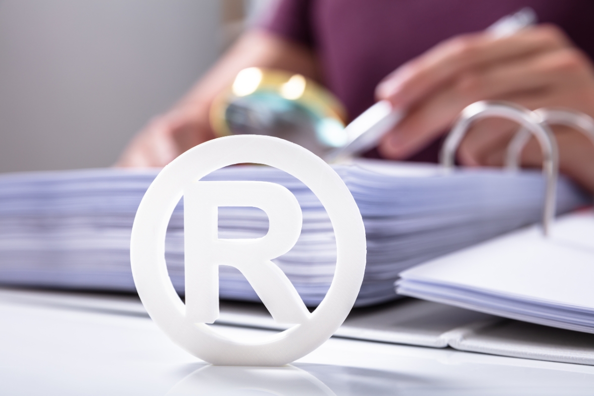 A registered trademark symbol in the foreground with blurred office paperwork and a person in the background.