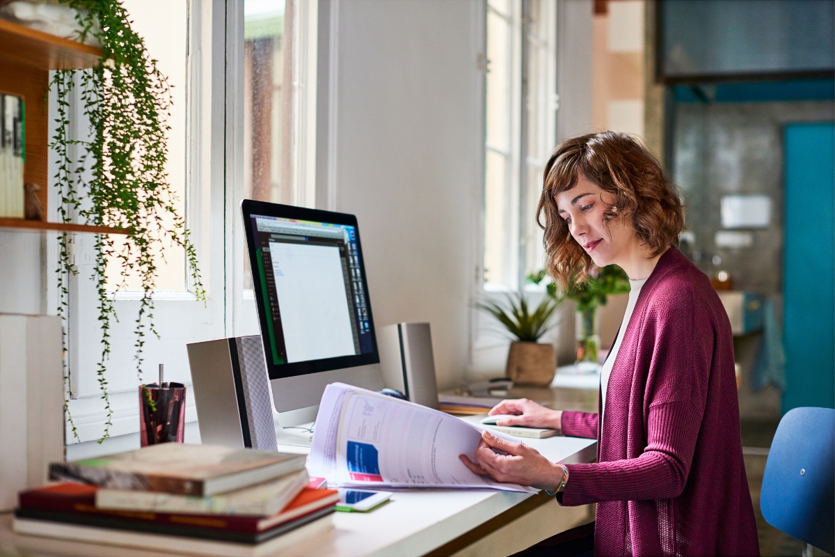 A woman reviews a document related to service-based business marketing while standing at her workstation with a computer monitor in the background.