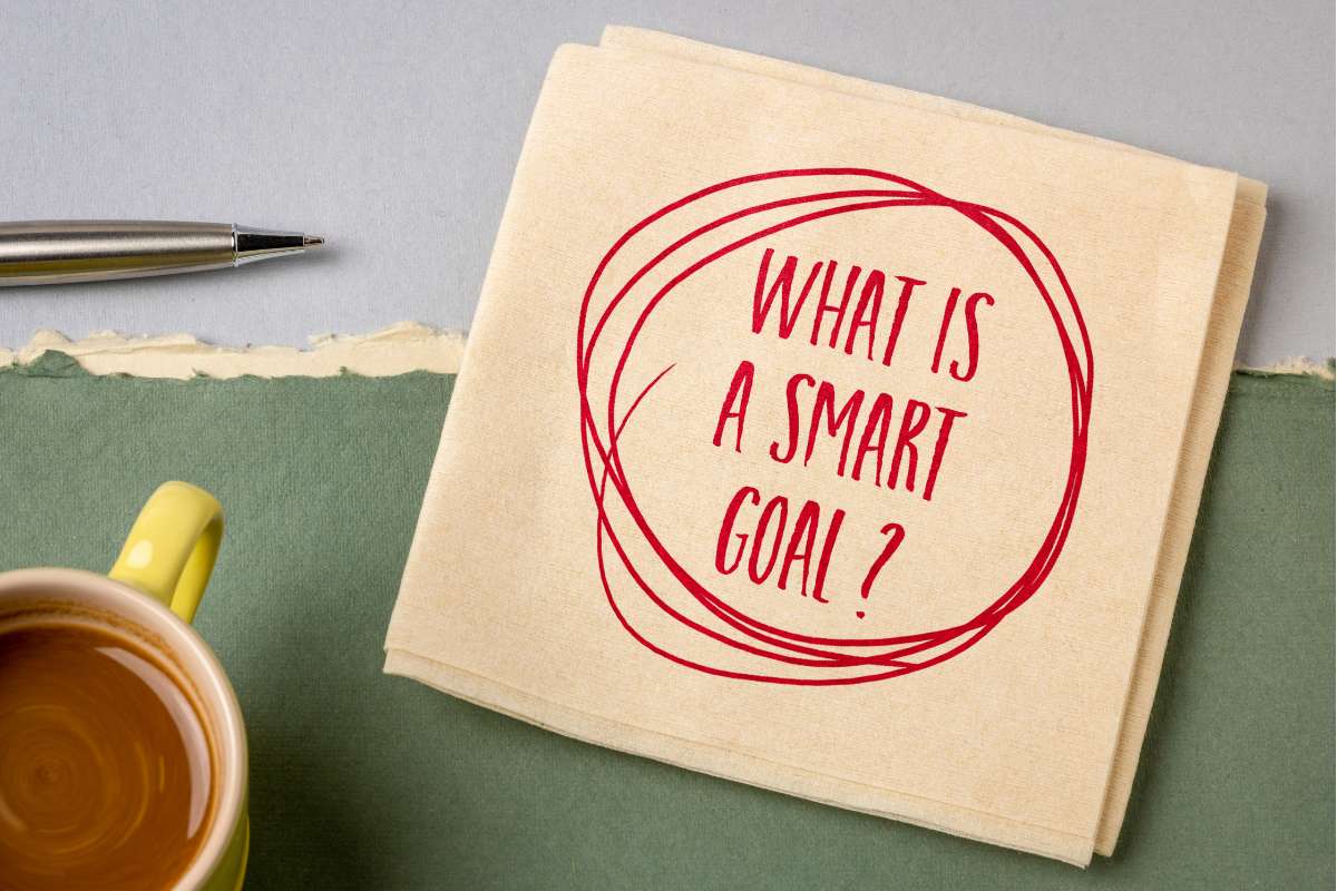 A note with "what is a smart goal?" written on it, alongside a pen and a cup of tea on a table.