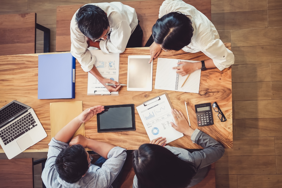 Four individuals collaborating on small business marketing ideas at a wooden table with laptops, tablets, documents, and a calculator.