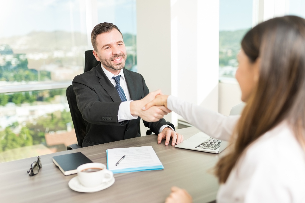 A professional man in a suit shakes hands with a woman across a desk in a bright office, focusing on customer relationship management, with documents and a coffee cup on the table.