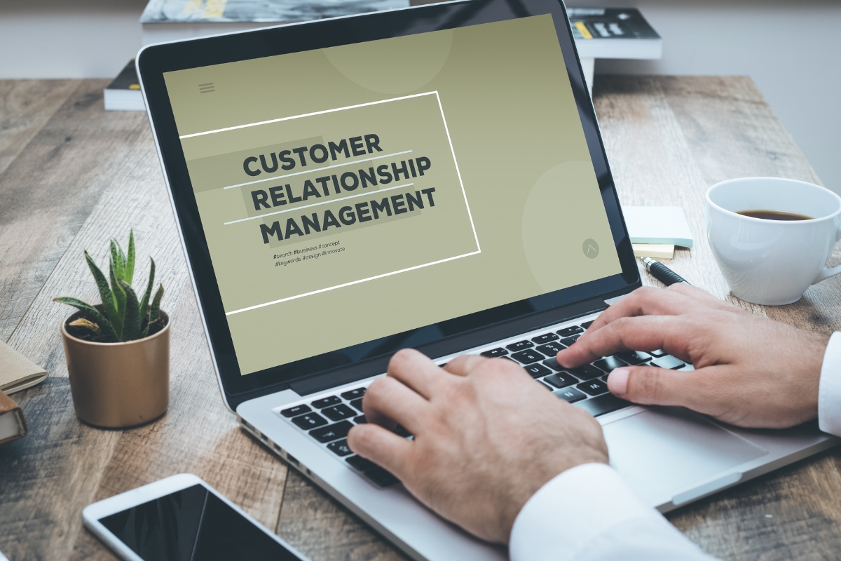 Close-up of a person using a laptop displaying "customer relationship management" on screen, with a coffee cup and plant beside it on a wooden desk.