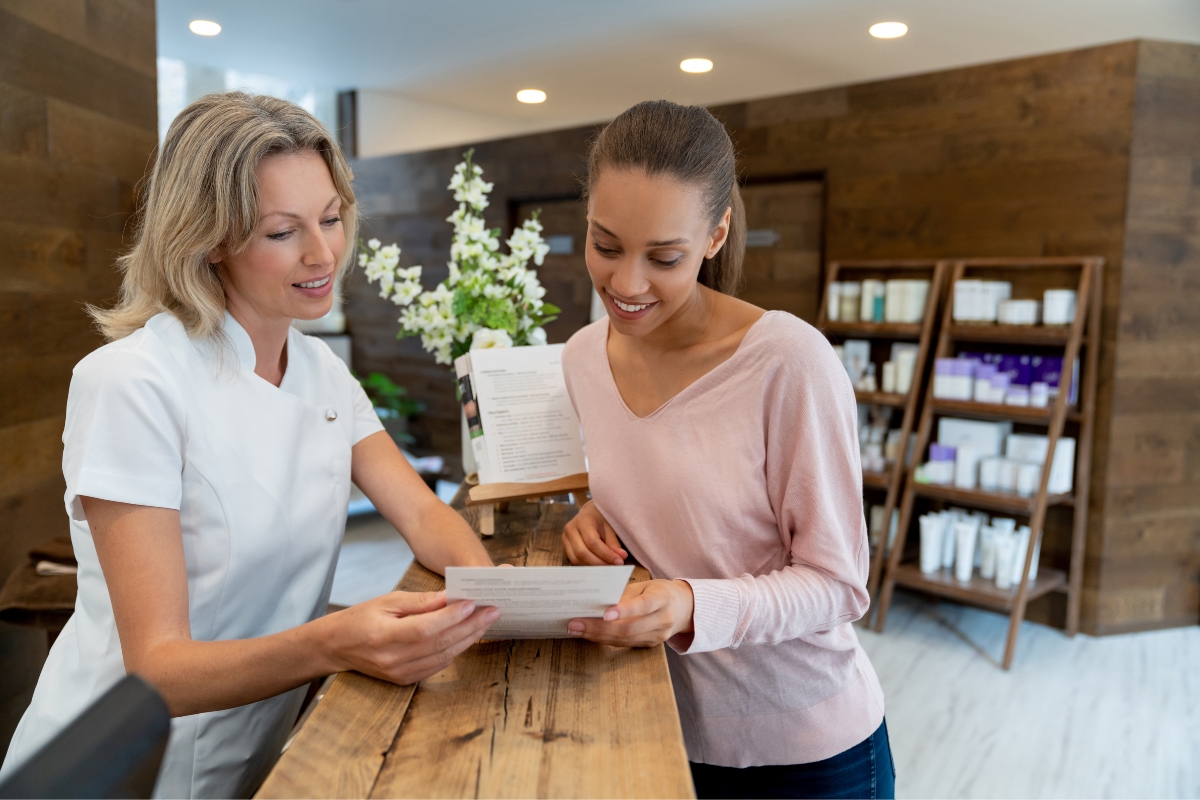 Two women, one in a white uniform, reviewing a customer relationship management document together at a spa reception desk with shelves of products in the background.