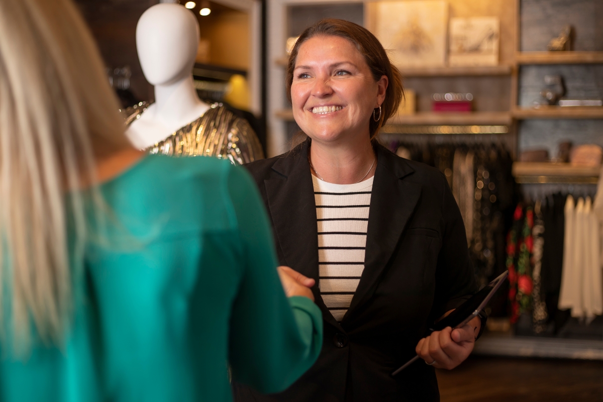 A smiling woman in a black blazer and striped shirt discussing customer relationship management with another woman in a boutique with clothes and a mannequin in the background.